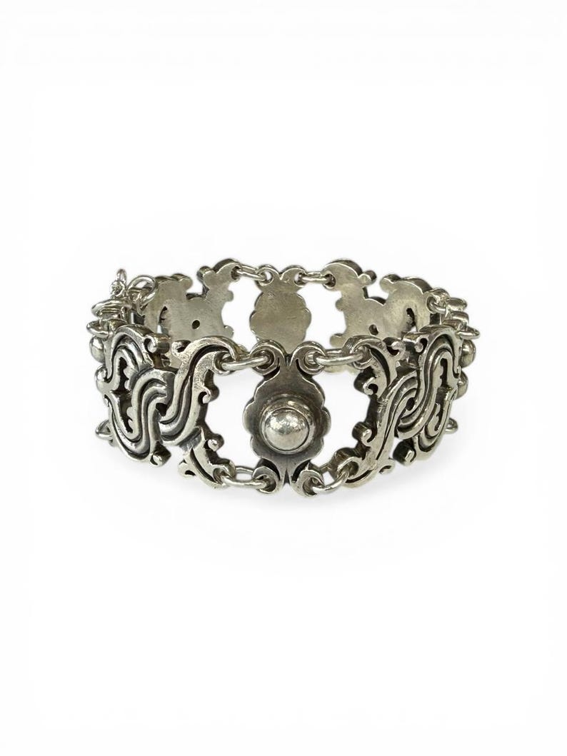 K&ouml;nnte beinhalten: Ein silberfarbenes Armband mit einem verzierten, durchbrochenen Design. Das Armband weist sich wiederholende Blumen- und Scrollmuster auf, die durch kleine Kettenglieder verbunden sind. Ein zentrales bl&uuml;tenf&ouml;rmiges Element mit erhabenem Mittelpunkt f&uuml;gt Details hinzu.