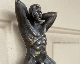 Può includere: Una scultura in bronzo di una figura maschile nuda con le braccia alzate sopra la testa. La figura ha una corporatura muscolosa e un'espressione facciale dettagliata.