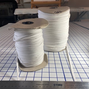 May include: Two spools of white elastic cord are on a grid-patterned surface. A metal ruler with centimetre measurements is in the foreground. The larger spool has the number "350" written on it. The background includes a painting and a sewing table.