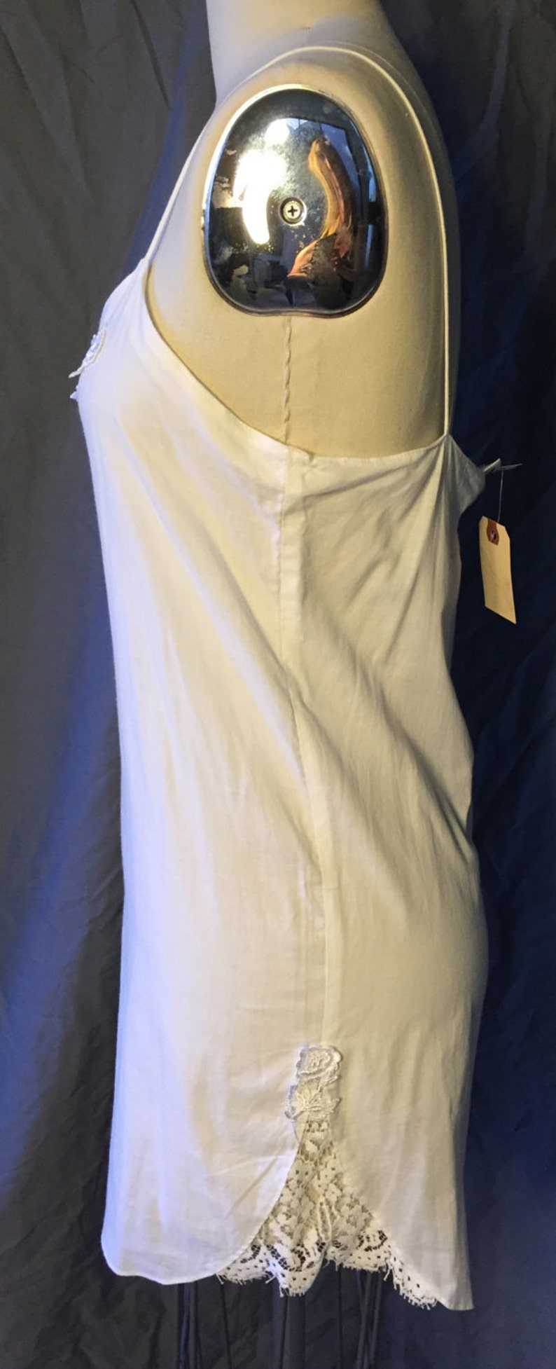 K&ouml;nnte beinhalten: Wei&szlig;es, &auml;rmelloses Schlupfkleid mit einem Spitzenbesatz am Saum. Das Kleid befindet sich auf einer wei&szlig;en Kleiderpuppe.