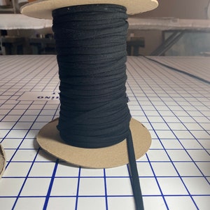 May include: A spool of black elastic cord wound around a cardboard tube. The cord is lying on a white surface with blue grid lines.
