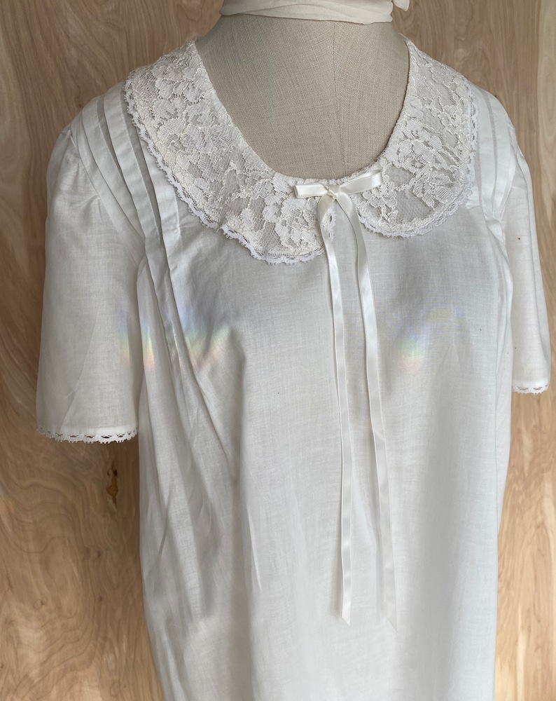 May include: White cotton nightgown with a lace collar and a satin bow. The nightgown has short sleeves and a gathered bodice.
