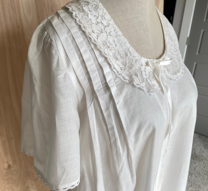 May include: White cotton nightgown with a lace collar and a bow at the neckline. The nightgown has a pleated bodice and short sleeves.