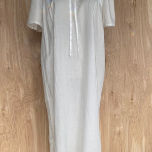 May include: A white cotton nightgown with a lace collar and a satin bow. The nightgown has short sleeves and a lace trim around the bottom.