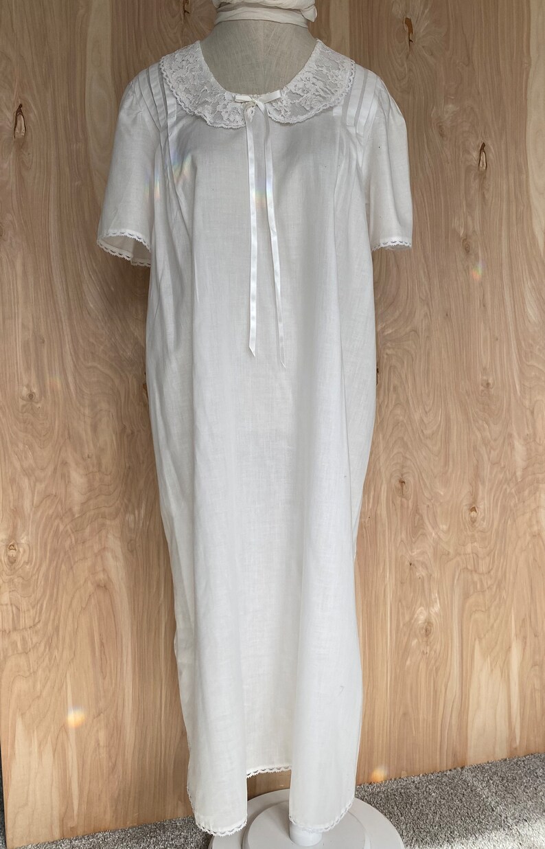 May include: A white cotton nightgown with a lace collar and a satin bow. The nightgown has short sleeves and a lace trim around the bottom.