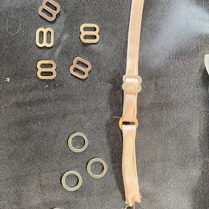 Pode incluir: Uma coleção de materiais para fazer sutiãs, incluindo vários deslizadores de metal, anéis e um pedaço de material de alça cor de rosa com um ajustador de metal. Um alfinete amarelo prende a extremidade da alça.