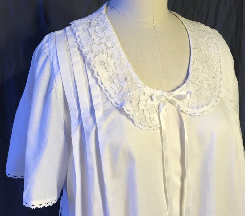 May include: White cotton nightgown with a lace collar and a satin bow. The nightgown has short sleeves and a gathered skirt.