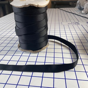 Op de afbeelding: Een rol zwart elastisch lint op een wit geblokt oppervlak. Het lint is om een kartonnen kern gewikkeld.