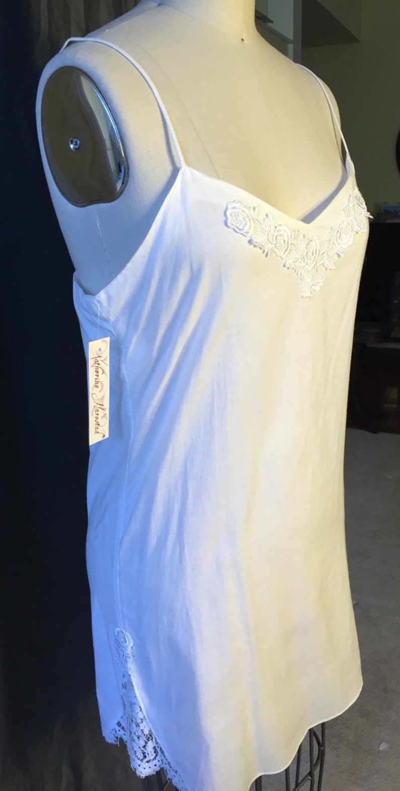 K&ouml;nnte beinhalten: Wei&szlig;es Schlupfkleid mit zarter Spitzenborte und Blumenstickerei am Ausschnitt. Das Kleid befindet sich auf einer wei&szlig;en Puppe. Das Etikett lautet "Katherine Hamnett".