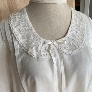 May include: A white cotton blouse with a lace collar and a white satin bow. The blouse has short sleeves and a relaxed fit.