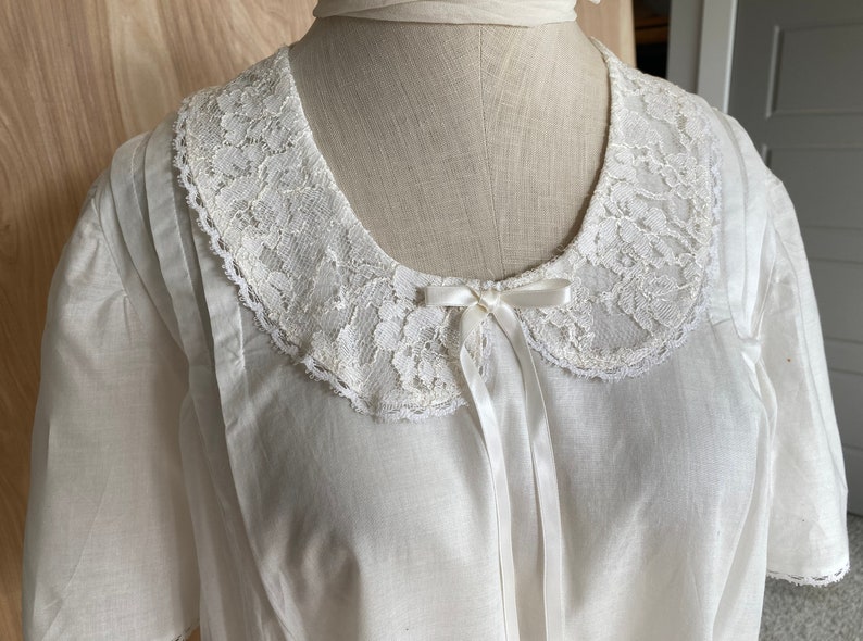 May include: A white cotton blouse with a lace collar and a white satin bow. The blouse has short sleeves and a relaxed fit.