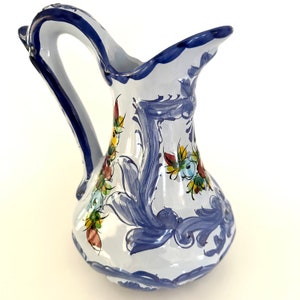 May include: A white ceramic pitcher with a blue handle and floral designs in blue, yellow, red, and green.