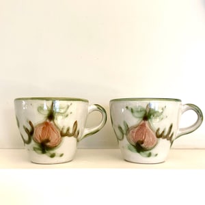 May include: Two ceramic mugs with a white base and green trim. Each mug features a hand-painted design of a red onion with green leaves and brown stems. The mugs have small handles and are set against a white background.