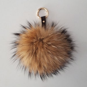 Può includere: Un portachiavi con pompon in pelliccia marrone e nero con un anello in metallo dorato e un cinturino in pelle marrone.