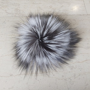 May include: A round, fluffy pom-pom made of black and white faux fur. The pom-pom has a dense, solid form, with long, soft strands extending outwards. The contrasting colours create a striking visual effect, suitable for craft projects or fashion accessories.