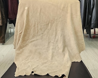 May include: A large, irregular piece of beige suede leather with a soft, matte texture and natural, unfinished edges. The material is draped over a dark surface, showcasing its flexible, pliable nature and consistent light tan coloration throughout.