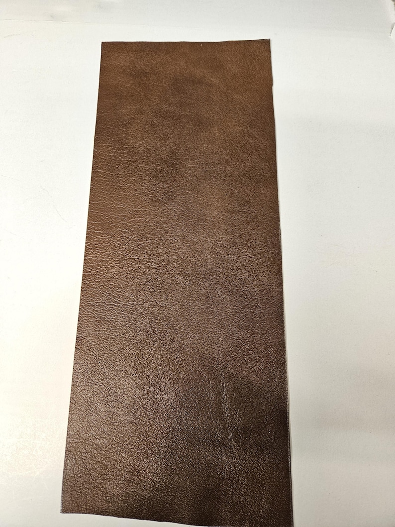 May include: A rectangular piece of brown leather with a textured surface. The leather is a rich, warm brown color, and the texture appears to be a fine grain. The piece is likely intended for use in crafting or other projects.