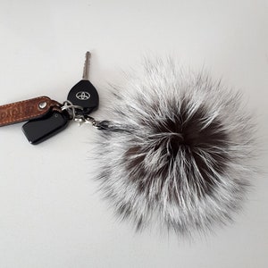 May include: A fluffy white and brown fur pom pom keychain attached to a silver key ring with a black key fob and a brown leather strap with the word "Toyota" embossed on it.