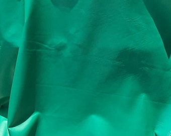May include: A large, rectangular piece of smooth, vibrant green leather draped over a chair. The material has a consistent, solid emerald green color with a soft sheen and natural folds that create subtle shadows across the surface of the hide.