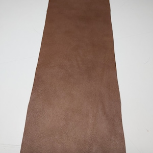 May include: A rectangular piece of brown leather with a textured surface. The leather is a solid, warm brown color and appears to be a single piece. The edges are slightly uneven, suggesting it may be cut from a larger hide.