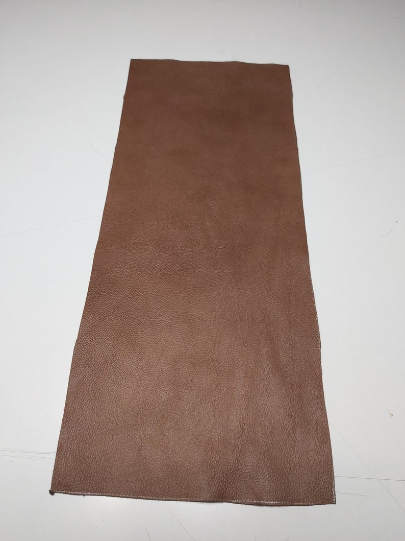 May include: A rectangular piece of brown leather with a textured surface. The leather is a solid, warm brown color and appears to be a single piece. The edges are slightly uneven, suggesting it may be cut from a larger hide.