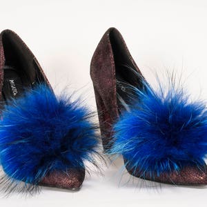 Puede incluir: Un par de zapatos de tacón alto de color rojo oscuro con pompones de piel azul unidos a los dedos. Los zapatos tienen un acabado brillante y el nombre de la marca "Migato" es visible en el zapato izquierdo.
