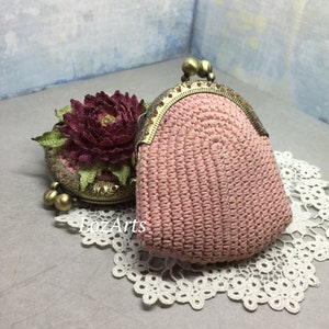 May include: A pink crocheted coin purse with a brass clasp and decorative beads. The purse is sitting on a white lace doily.