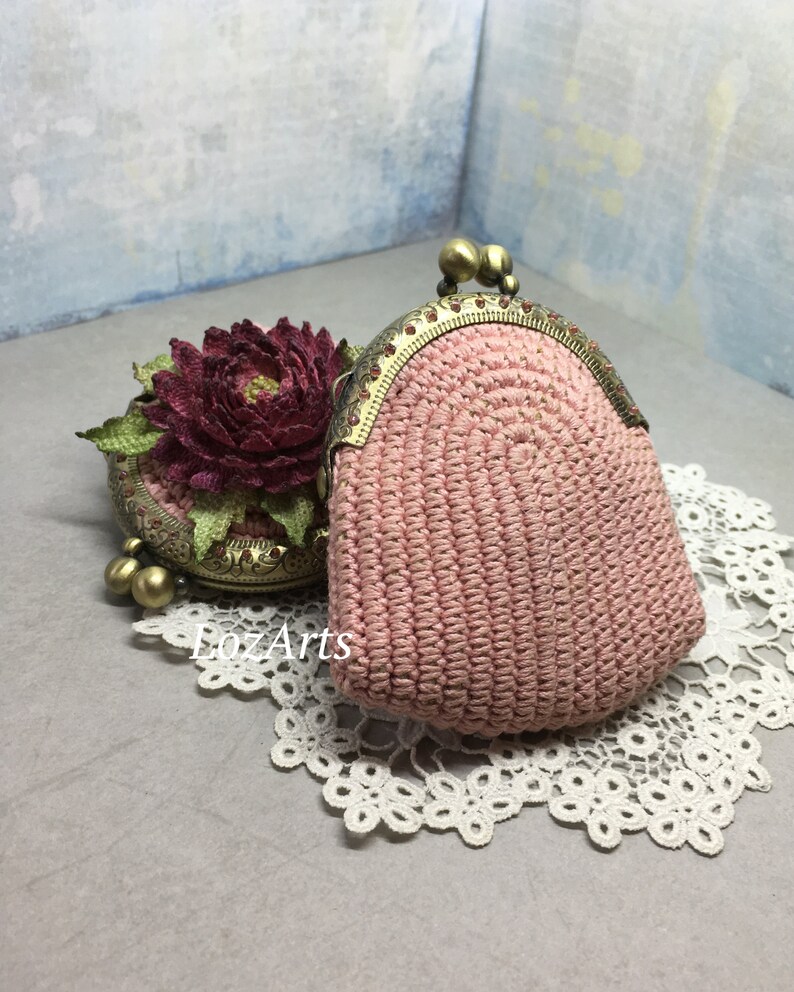 May include: A pink crocheted coin purse with a brass clasp and decorative beads. The purse is sitting on a white lace doily.