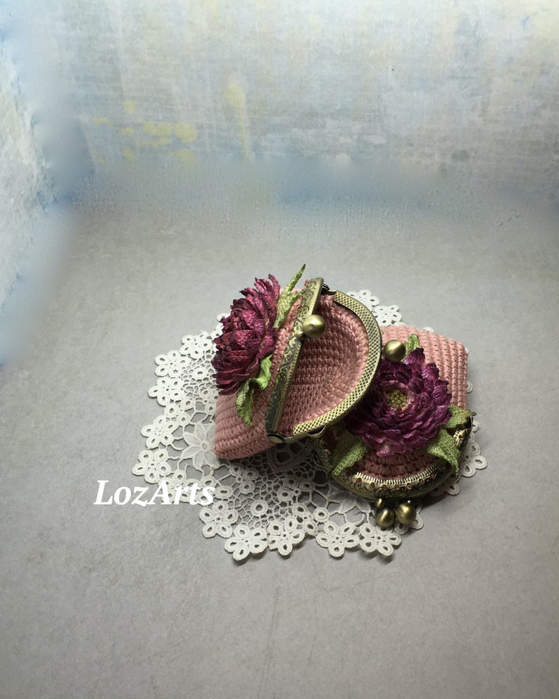 May include: Two pink crocheted coin purses with floral embellishments and gold metal clasps. The purses are on a white doily.