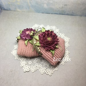 May include: A pink crocheted coin purse with a floral design and a metal clasp. The purse is decorated with two pink fabric flowers.
