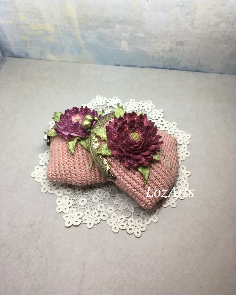 May include: A pink crocheted coin purse with a floral design and a metal clasp. The purse is decorated with two pink fabric flowers.