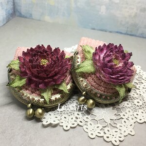 May include: Two crocheted coin purses with floral embellishments. The purses are pink with a burgundy flower and green leaves. The purses have a brass clasp and are sitting on a white lace doily.