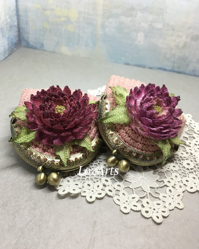 May include: Two crocheted coin purses with floral embellishments. The purses are pink with a burgundy flower and green leaves. The purses have a brass clasp and are sitting on a white lace doily.