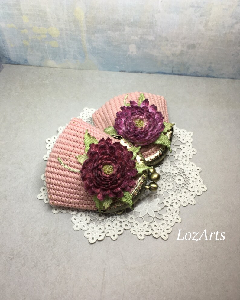 May include: Two pink crocheted coin purses with floral embellishments. The purses have a vintage-style clasp and are sitting on a white lace doily.