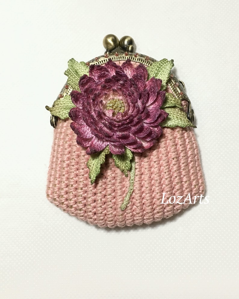 May include: A pink crocheted coin purse with a pink and green fabric flower and a brass clasp. The purse has the text "LozArts" on the bottom.
