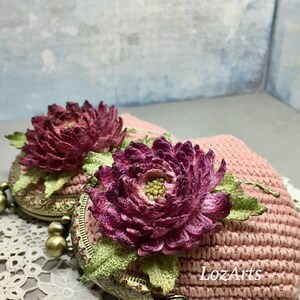 May include: A pink crocheted coin purse with a floral design. The purse has a metal clasp and is decorated with two large, purple flowers.