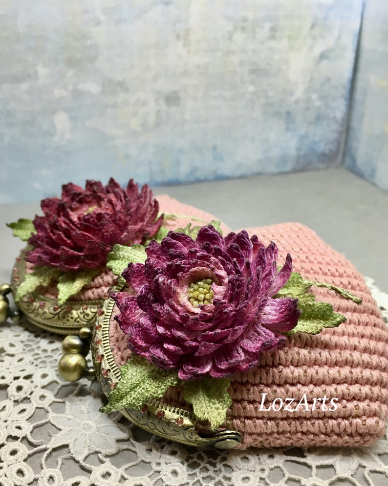 May include: A pink crocheted coin purse with a floral design. The purse has a metal clasp and is decorated with two large, purple flowers.