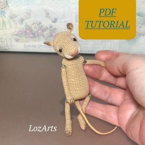 Puede incluir: Tutorial PDF para un patrón de ratón amigurumi de crochet. El patrón incluye instrucciones para tejer un pequeño ratón beige con una cola larga. El ratón está sentado en una mano.