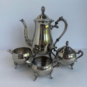 Puede incluir: Un juego de té plateado con detalles ornamentados. El juego incluye una tetera, un azucarero con tapa y dos cremeras. La tetera tiene un pico curvo y un asa decorativa. Las piezas son reflectantes y están sobre una superficie blanca.