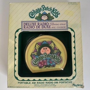 May include: A yellow and green box containing a Cabbage Patch Kids Deluxe Radio. The radio is yellow with a Cabbage Patch Kid face on it. The box states "Deluxe Radio with Carrying Strap" and "Portable AM Radio".
