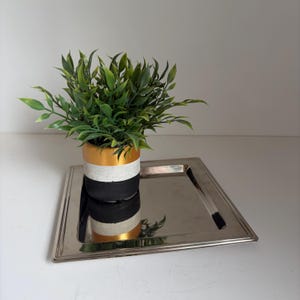 Puede incluir: Una bandeja cuadrada de metal plateado pulido que sostiene una maceta cilíndrica. La maceta presenta tres bandas horizontales de color dorado, blanco y negro, y contiene una exuberante planta verde artificial con hojas puntiagudas y tallos visibles.
