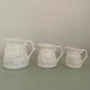 Puede incluir: Conjunto de tres jarras de cerámica blanca con un diseño de vid. Las jarras son de diferentes tamaños, con la jarra más grande en la parte posterior y la más pequeña en la parte delantera.