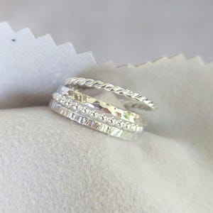 May include: A set of four silver stacking rings. The rings are made of different textures, including a twisted band, a hammered band, and a beaded band.