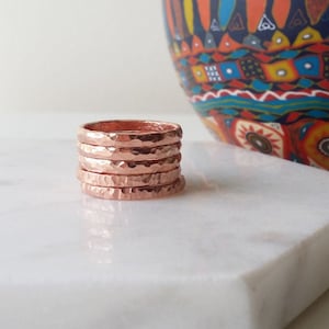May include: Six hammered copper rings stacked on a white marble surface. The rings are a warm rose gold colour with a textured, faceted surface. A colourful decorative bowl is in the background.