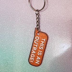 May include: Orange keychain with the text "THIS IS AN OUTRAGE!" in white. The keychain has a silver chain and ring. The background is a gradient of pink, blue, and white.