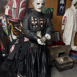 Może przedstawiać: Figurka Pinheada w naturalnej wielkości z serii filmów Hellraiser. Figurka ma bladą twarz z szpilkami, czarną kurtkę ze skóry i czarną spódnicę. Figurka trzyma pudełko z puzzlami. Inne przedmioty w tle to torba golfowa i grafika.