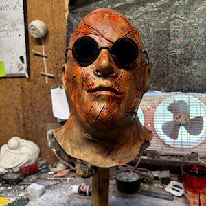 May include: A detailed, weathered mask with a realistic human face, featuring dark round sunglasses and numerous red gashes. The mask is mounted on a wooden stand, showcasing a horror or Halloween theme.