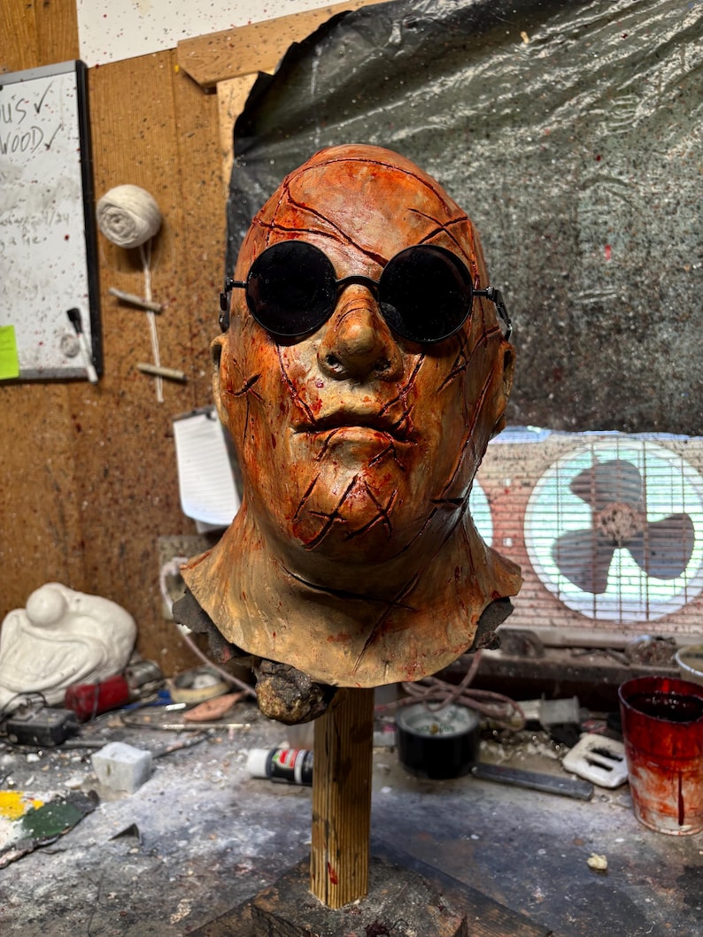 May include: A detailed, weathered mask with a realistic human face, featuring dark round sunglasses and numerous red gashes. The mask is mounted on a wooden stand, showcasing a horror or Halloween theme.