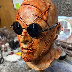 May include: A detailed, orange-toned mask with deep red cuts and black round sunglasses. The mask is mounted on a wooden dowel, suggesting a display or prop. The overall aesthetic is reminiscent of horror or Halloween.