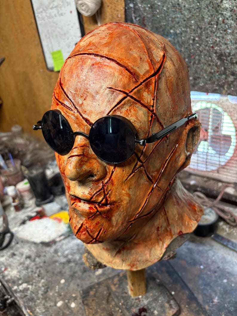 May include: A detailed, orange-toned mask with deep red cuts and black round sunglasses. The mask is mounted on a wooden dowel, suggesting a display or prop. The overall aesthetic is reminiscent of horror or Halloween.